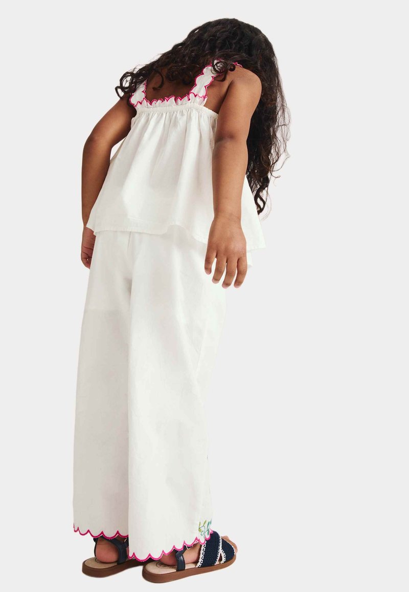 Niña joven con cabello largo y rizado, vestida con una blusa sin mangas blanca y pantalones anchos con bordes rosados en forma de ondas, de pie de espaldas.