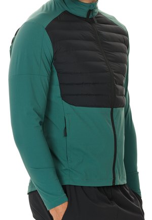 Uomo che indossa una giacca a zip teal e nera con pannello imbottito sul petto e tasche laterali, in piedi con le mani nelle tasche dei pantaloni neri.