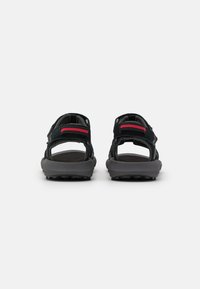 Sandalias negras con correas ajustables, suela gris y suela texturizada. Presentan un acento rojo en la correa del talón y un diseño abierto.