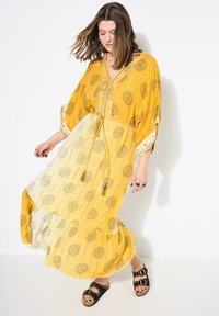 Maxi dress gialla con motivi floreali, vita cinchata e frange. Sezione inferiore trasparente, maniche a tre quarti con bordo decorativo.