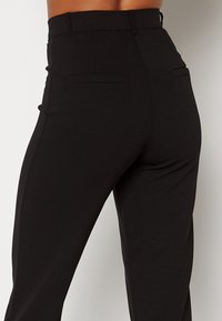 Pantalon noir taille haute à texture lisse, avec deux poches arrière et une coupe effilée aux jambes. Lignes épurées et détails minimalistes.