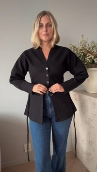 Casaco preto de mangas longas com decote em V, fecho de cinco botões, laços laterais e detalhes costurados, combinado com jeans azuis. Textura suave.