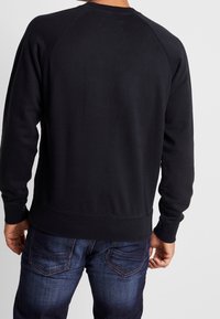 Persona che indossa un semplice felpa nera e jeans blu scuri, vista da dietro su uno sfondo chiaro e neutro.