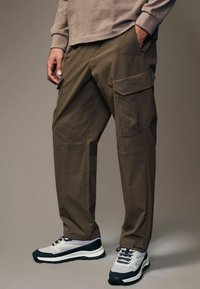 Pantalones cargo de tejido marrón apagado con un ajuste relajado, que cuentan con múltiples bolsillos y una textura suave. Combinados con zapatillas grises y negras.