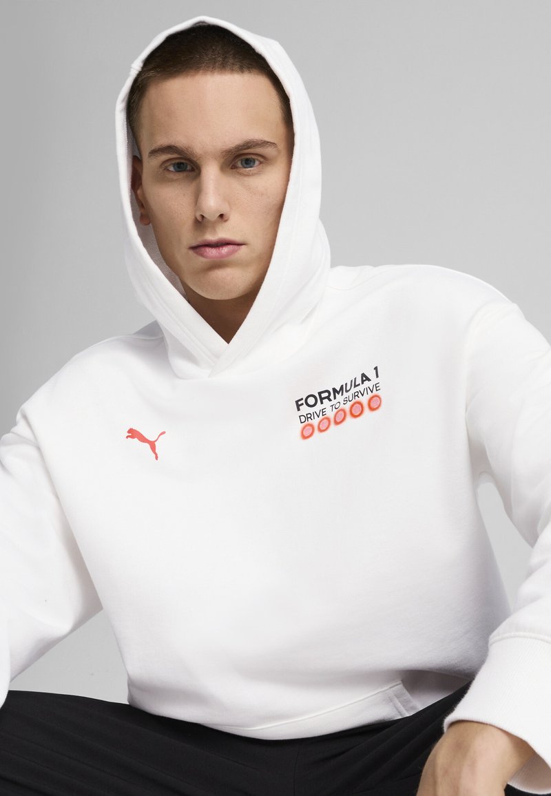 Fehér kapucnis pulóver elülső kenguruzsebbel, fekete és narancssárga szöveggel "FORMULA 1" és "DRIVE TO SURVIVE", egy kis piros Puma logóval.