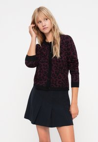 Black and burgundy leopard print cardigan with long sleeves, round neck, and button closure. Styled with a black pleated skirt.