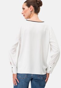 Weiße Bluse mit langen Ärmeln, runder Ausschnitt und schwarzen Akzenten. Mit Knopfdetails an den Ärmelöffnungen. Kombiniert mit blauen Jeans.
