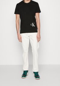 Camiseta de algodón negra con cuello redondo, que presenta un logo blanco de "CK" en la parte inferior izquierda. Combinada con vaqueros blancos y zapatillas de deporte en color teal.