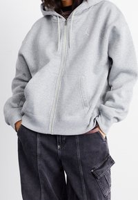 Sudadera gris con cremallera, bolsillo canguro, capucha con cordón y detalle de logo. Textura suave y corte holgado, combinada con pantalones cargo oscuros.