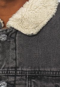 Zwarte spijkerjas met crèmekleurige nep bontkraag. Voorzien van metalen knopen en zichtbare stiksels langs de naden. Gestructureerde stof.