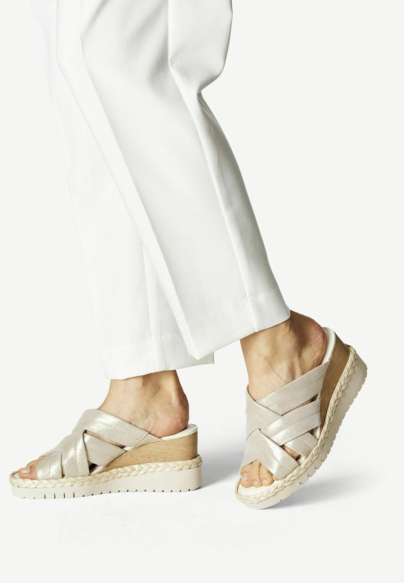 Pieds portant des sandales compensées beiges avec des brides croisées, assorties à un pantalon blanc sur un fond clair uni.
