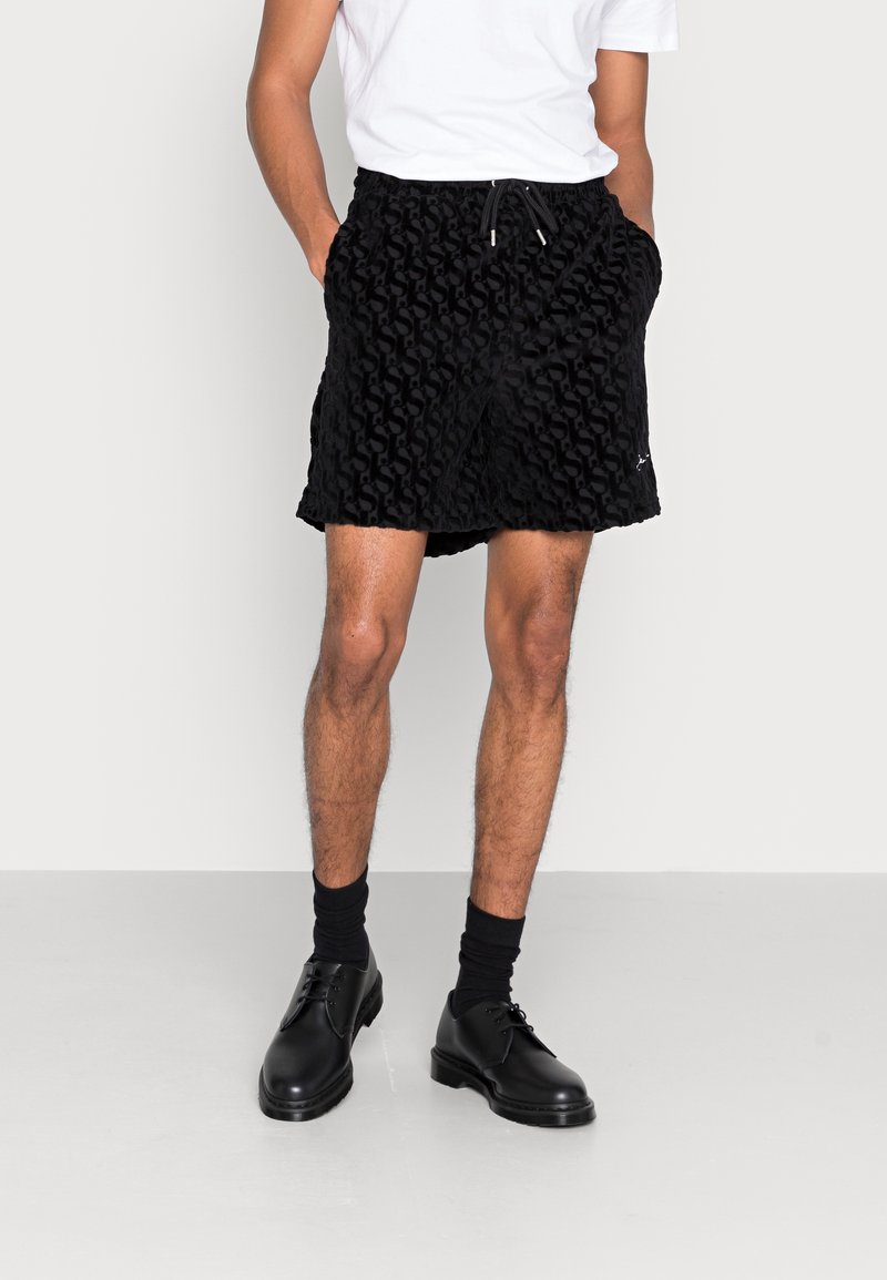 Shorts noirs texturés avec un motif ondulé, dotés d'une taille élastique et d'un cordon de serrage ajustable. Associés à des chaussures habillées noires et des chaussettes.