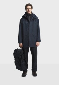 Marinblå vattentät parka med huva, tryckknappar och två framfickor; kombinerat med svarta byxor och en svart ryggsäck.