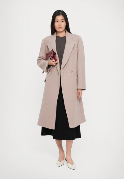 Woman wearing a beige long coat over a dark dress, holding a red clutch, and wearing white pointed-toe shoes, standing against white background.