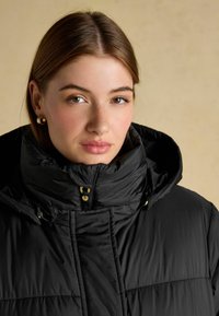 Černá péřová bunda s vysokým límcem, zlatými doplňky a prošívanou strukturou. Tkanina vypadá hladce a mírně leskle.