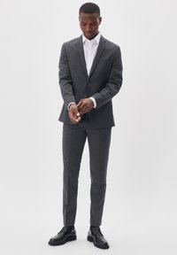 Traje gris con solapa muesca, cierre de un solo botón y corte ajustado. Combinado con una camisa blanca y zapatos de cuero negros. Textura de tela suave.