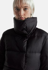 Černá péřová bunda s vysokým, ohnutým límcem, prošívaným designem a hladkým povrchem. Disponuje lesklým povrchem a přiléhavým střihem.