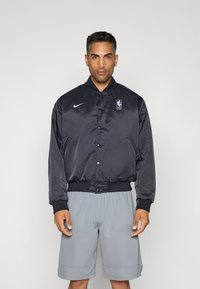 Mies, joka pukee yllensä mustan satiinin NBA-takin, jossa on violetit logot, sekä harmaat koripallosortsit vaaleaa taustaa vasten.