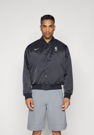 Mand iført en sort satin NBA-jakke med lilla logoer og grå basketballshorts foran en ensfarvet lys baggrund.