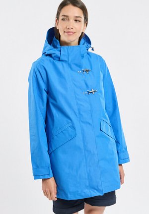 Femme portant un imperméable à capuche bleu vif avec deux fermetures à boutons à bascule et des poches à rabat, regardant légèrement vers sa gauche.