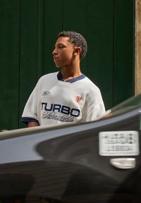 White short-sleeve jersey with blue accents, featuring "TURBO" text and a logo, worn by a person beside a dark vehicle and green background.