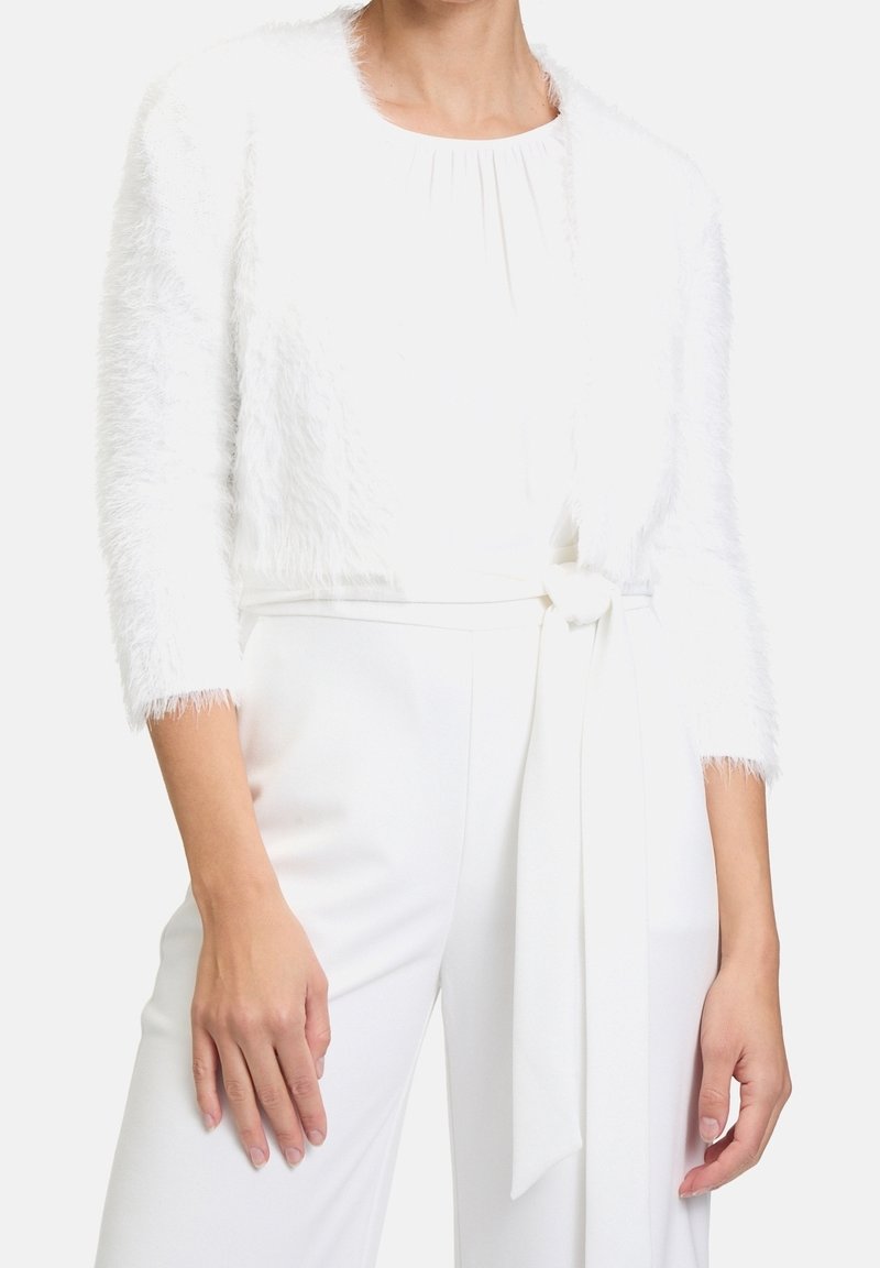 Vit faux fur bolerojacka med trekvartslånga ärmar, mjuk textur, öppen framsidan design och en kort silhuett, bärs över en vit outfit.