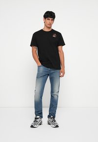 T-shirt en coton noir avec un petit graphique d'araignée sur la poitrine gauche, associé à un jean en denim bleu clair et des baskets blanches avec des accents noirs.