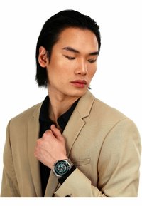 Silberne Armbanduhr mit grünem Ziffernblatt, Metallarmband und mehreren Ziffernblättern. Getragen mit einem beigen Blazer über einem schwarzen Hemd.