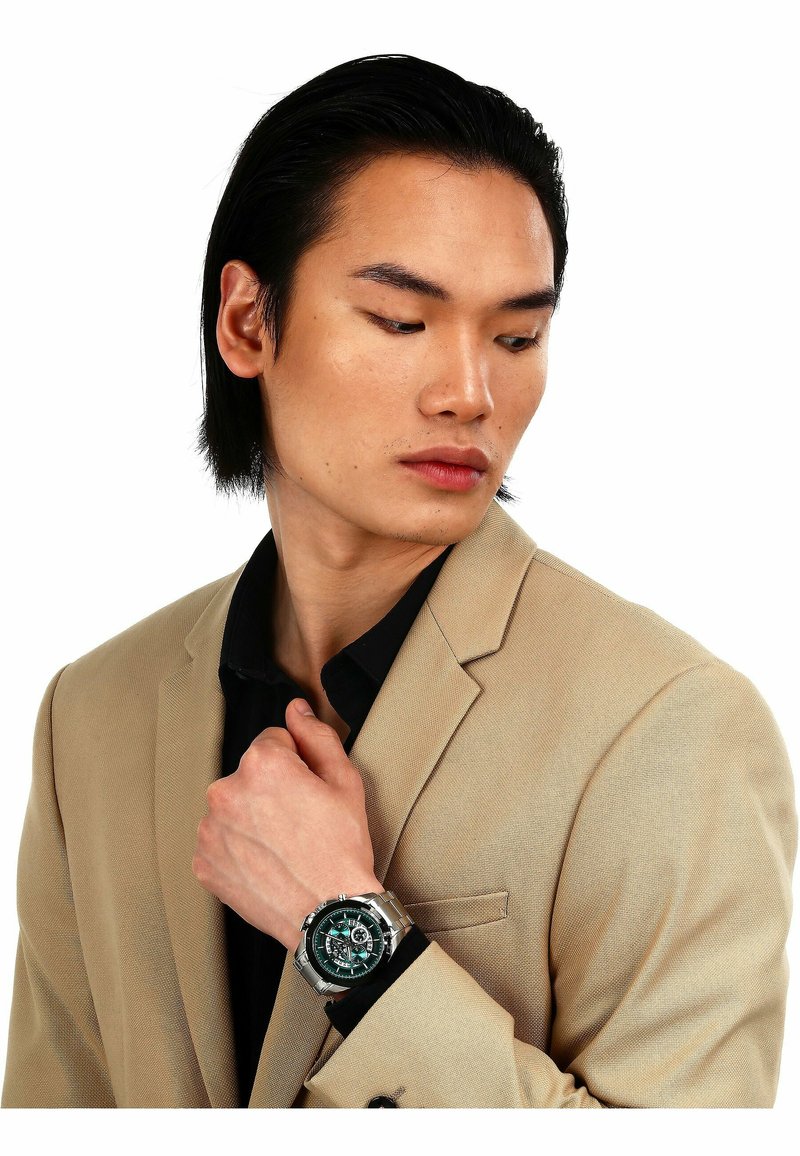 Silberne Armbanduhr mit grünem Ziffernblatt, Metallarmband und mehreren Ziffernblättern. Getragen mit einem beigen Blazer über einem schwarzen Hemd.