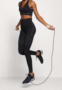 Sujetador deportivo negro con un frente cruzado, mallas negras de cintura alta con acentos texturizados y zapatillas deportivas negras con suelas blancas.