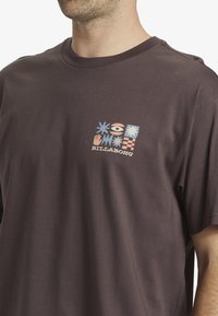 Camiseta de algodón marrón con un diseño gráfico multicolor que presenta formas y símbolos en la parte izquierda del pecho. Cuello redondo clásico y mangas cortas.