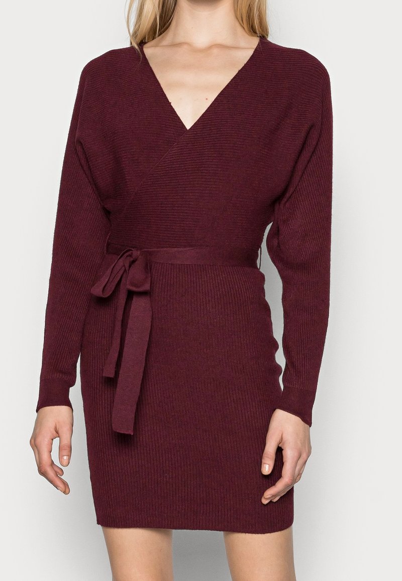 Femme portant une robe portefeuille bordeaux à manches longues côtelées avec une ceinture nouée à la taille, vue du cou jusqu'au milieu de la cuisse.