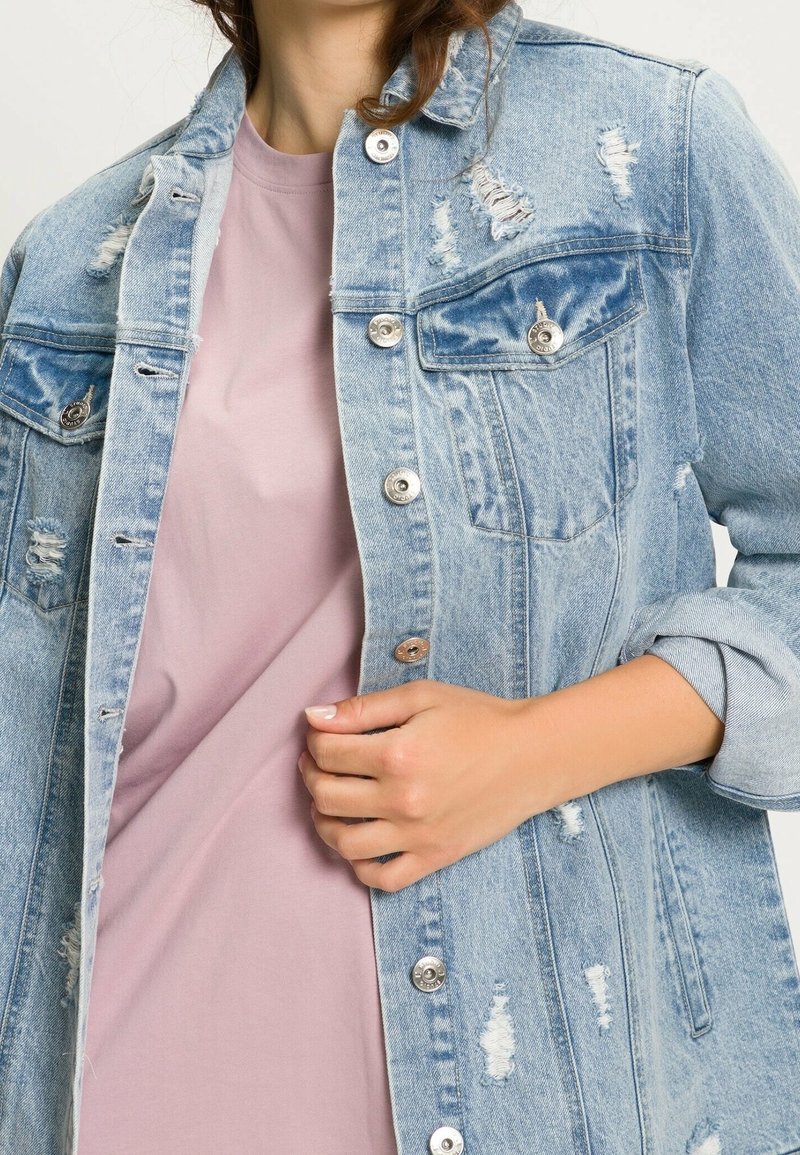 Ljusblå distressed denimjacka med silverknappar och fickor, lager på lager över en ljusrosa skjorta. Har uppvikta ärmar och trasiga kanter.