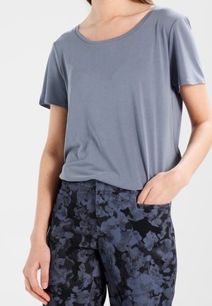 Person trägt ein graues kurzärmeliges Hemd und schwarze Hose mit blauem Blumenmuster, eine Hand in der Tasche vor weißem Hintergrund.