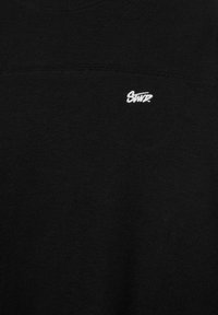 Tissu noir avec un petit texte blanc brodé "Swvz" près du centre supérieur, montrant les détails de la texture et des coutures.