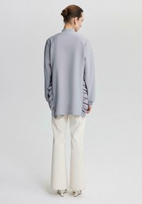 Top lung gri cu mâneci lungi și detalii pliate pe părți, purtat cu pantaloni largi de culoare crem și tocuri albe ascuțite, pe un fundal neutru.