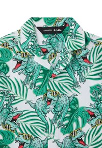 Camicia in cotone a maniche corte con sfondo bianco e illustrazioni di dinosauri verdi con occhiali da sole, immerse in motivi di foglie tropicali.