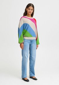 Pull surdimensionné coloré avec de larges rayures diagonales rose, bleu, vert et beige, associé à un jean droit bleu clair et des ballerines noires.