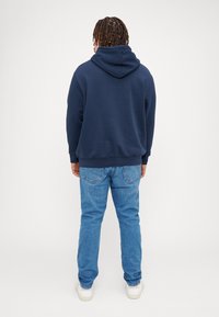 Marineblå hættetrøje med kengurulomme, ribbede manchetter og nederste kant. Kombineret med blå jeans og hvide sneakers, set fra bagsiden.