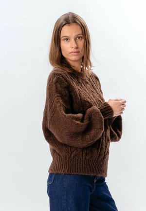 Pruunide õlgadeni juustega naine, kellel on peas jämeda palmiku mustriga pruuni värvi kampsun ja sinised teksad, seisab tavalise heleda tausta ees.