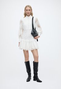 Γυναίκα που φοράει λευκό δαντελένιο φόρεμα με φουσκωτά μανίκια, μαύρες μπότες μέχρι το γόνατο και κρατά μαύρη τσάντα ώμου, στέκεται μπροστά σε λευκό φόντο.