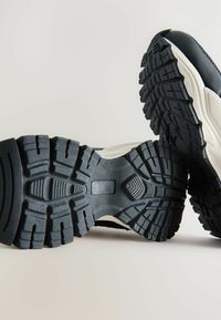 Talpă din cauciuc negru cu adâncimi, mijloc alb și partea superioară texturată. Are un design curbat pentru stabilitate și aderență pe diverse suprafețe.