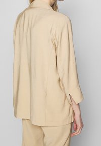 Beige, lightweight, collared blazer with a relaxed fit, featuring elbow-length sleeves and side slits for added movement.