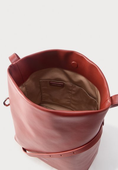 Sac seau en cuir brun rougeâtre ouvert, montrant une doublure intérieure beige avec une petite poche intérieure et une fermeture à pression magnétique.