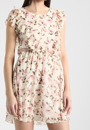 Kobieta w kremowej sukience bez rękawów z różowym kwiatowym wzorem i falbankami na wysokości klatki piersiowej i ramion.