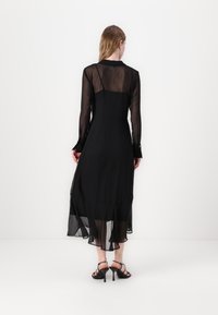 Svart långärmad klänning med genomskinlig överdel och flowande fåll. Har knäppning vid ärmsluten och en åtsittande bodice, kombinerad med svarta remsandaler.