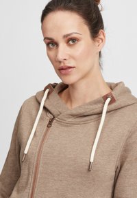 Lys brun hettegenser med glidelås, med snorhette, beige detaljer og en myk tekstur. Inkluderer bronsefarget beslag.