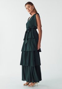Dunkelgrünes, mehrlagiges Maxikleid mit V-Ausschnitt und Taillengürtel, das aus weichem, fließendem Stoff besteht und kantige Saumdetails aufweist.