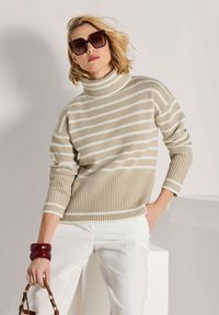 Gestreifter Rollkragenpullover in Beige und Weiß, gerippte Textur, mit Dreiviertelärmeln. Das Modell hält eine braune Handtasche und trägt übergroße Sonnenbrillen.