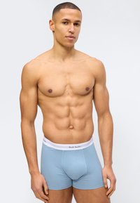 Homme en bonne forme avec des cheveux courts portant un boxer bleu clair à taille blanche marqué "Paul Smith" sur un fond uni.